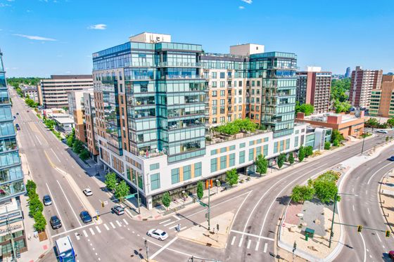 Modern luxury apartment building with sleek glass design and rooftop greenery, located at a vibrant city intersection with urban views.