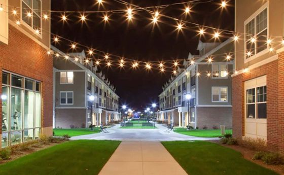 Charming apartment complex at night illuminated by string lights, showcasing a welcoming pathway and vibrant green lawns.
