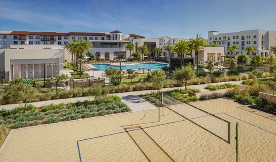 Luxurious apartment complex featuring a shimmering pool, sandy volleyball court, and lush landscaping under a clear blue sky.
