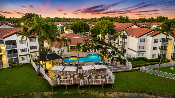 Captivating aerial view of a vibrant apartment community featuring lush greenery, a pristine pool area with sun loungers, and stylish outdoor seating under a stunning sunset sky.