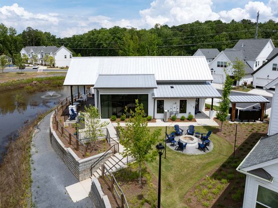Beautiful community center with a modern metal roof, overlooking a tranquil pond with a fountain and surrounded by lush greenery, featuring outdoor seating and a cozy fire pit area for relaxation.