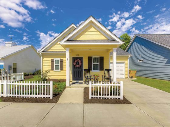 Charming yellow single-story house with a welcoming porch, rocking chairs, and a white picket fence under a bright, blue sky.
