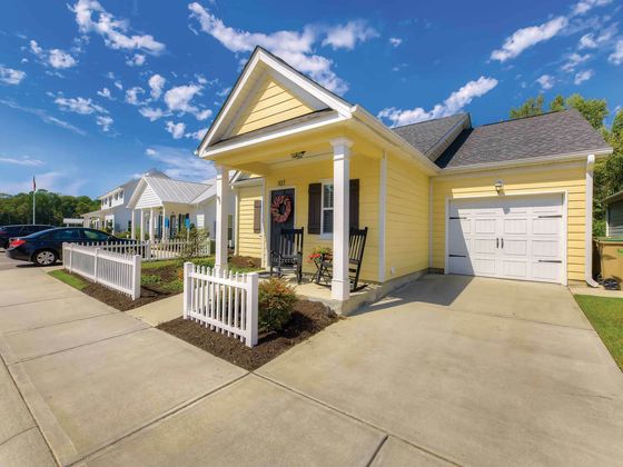 Charming yellow house with inviting porch, white picket fence, and lush surroundings under a bright blue sky.
