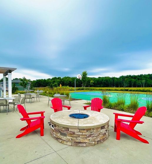 Vibrant outdoor area featuring a cozy stone fire pit with bright red chairs, overlooking a scenic basketball court and lush greenery.