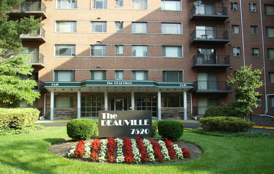 Charming brick apartment building with balconies and lush landscaping at The Deauville, featuring manicured gardens and a welcoming entrance.