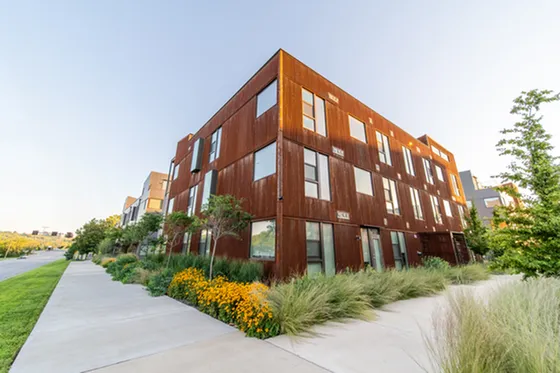 Modern apartment building with a unique rust-colored facade surrounded by lush greenery and vibrant yellow flowers on a sunny day.