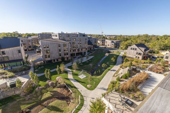 Scenic aerial view of modern wooden apartment complexes surrounded by lush greenery, pathways, and a peaceful stream.