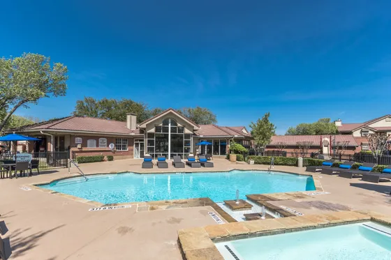 Luxurious apartment complex featuring a sparkling swimming pool with sun loungers, surrounded by lush landscaping and modern amenities under a clear blue sky.