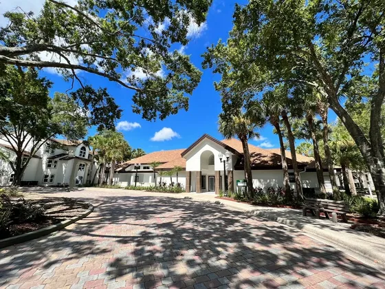Inviting apartment complex entrance with charming architecture, surrounded by lush greenery and a picturesque cobblestone pathway under a bright blue sky.