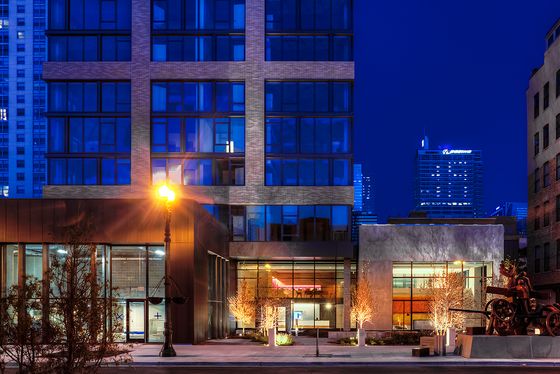 Modern urban apartment building with striking glass facade illuminated at night, surrounded by vibrant cityscape and warm street lighting.