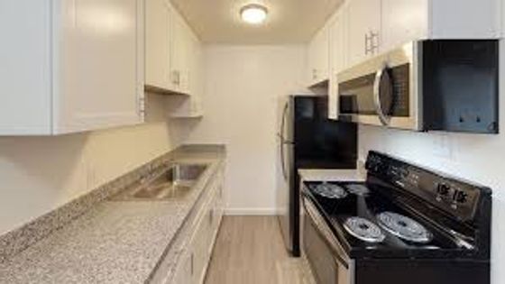 A long kitchen with white cabinets, gray granite countertops, a double sink, black appliances including a stove and refrigerator, and light wood flooring.