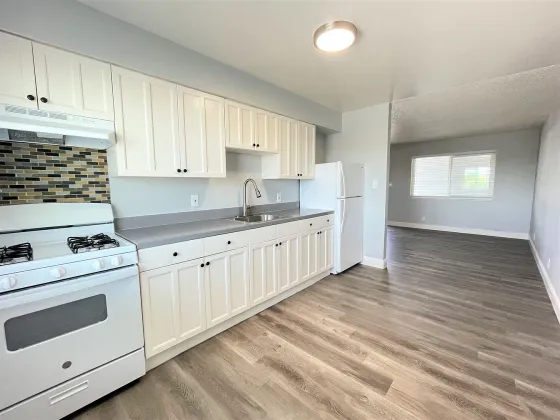 A modern kitchen with white cabinets, gray countertops, and a tiled backsplash. It includes a white stove, refrigerator, and stainless steel sink. Light wooden floor extends into an adjoining room with a window, creating an open, airy feel.