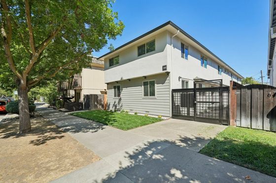 Two-story apartment building with white and grey exterior, large windows, and a gated wooden fence. A tree shades a sidewalk beside the lush green lawn. Bright, sunny day.