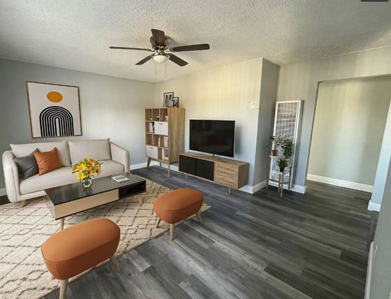 Modern living room with gray walls, featuring a beige couch with orange and gray cushions, a low table, artwork, TV on a stand, and two orange stools.