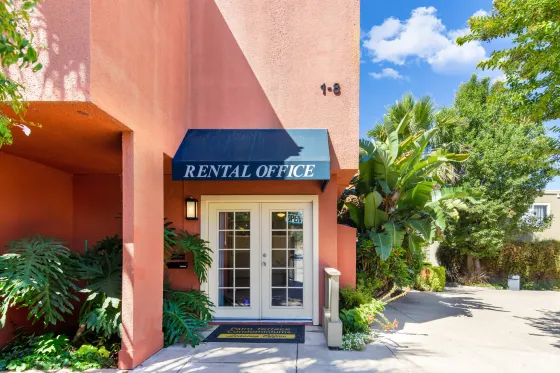 Entrance to a rental office with a blue awning and glass doors, surrounded by lush greenery. The building is peach-colored under a clear blue sky.