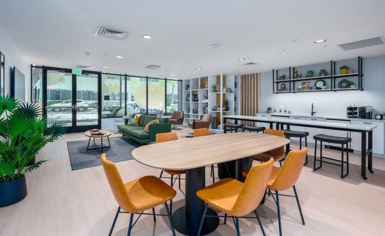 Modern lounge area with a large oval table and six orange chairs. Nearby, a green sofa and armchairs sit on a gray rug. The room is bright, with plants and a sleek kitchen.