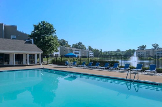 Inviting apartment complex pool area with lounge chairs, lush greenery, and a tree-lined backdrop under a clear blue sky.