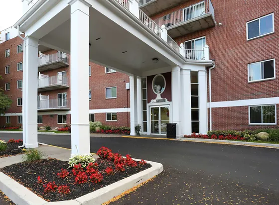 Elegant apartment entrance with red brick facade and landscaped gardens, featuring modern balconies and a welcoming drive-up area.