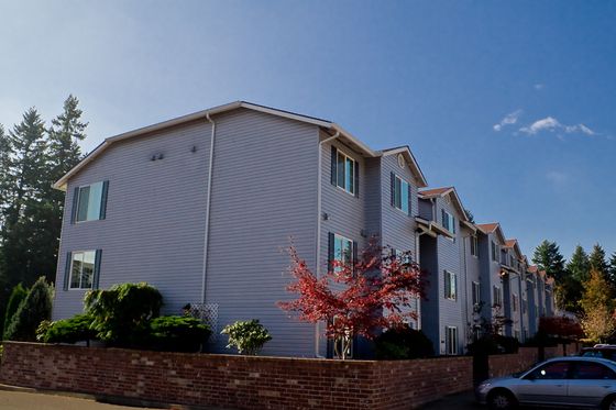Charming three-story residential building surrounded by vibrant trees and a welcoming brick wall, set against a clear blue sky.