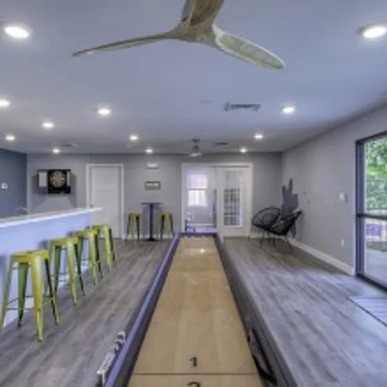Modern indoor community lounge at The Field Apartments in Springfield, MO, featuring a shuffleboard table, bar seating with lime green stools, contemporary lighting, and access to a patio through large glass doors.