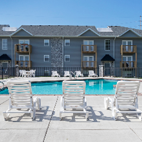 Four white pool lounge chairs lined up on a light-gray concrete deck facing a rectangular blue swimming pool, with a two-story beige and gray apartment building showing balconies and sliding-glass doors in the background under a clear sky at Concord Manor.