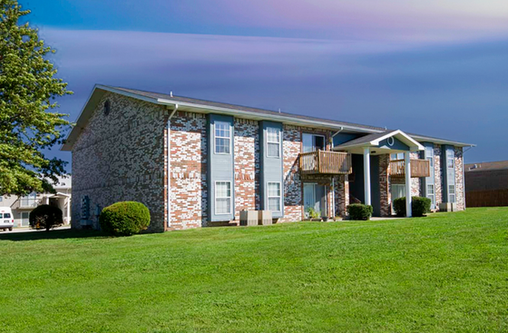 A brick apartment building with a gabled roof, surrounded by a lush green lawn. Wooden balconies and a clear blue sky enhance a serene atmosphere.