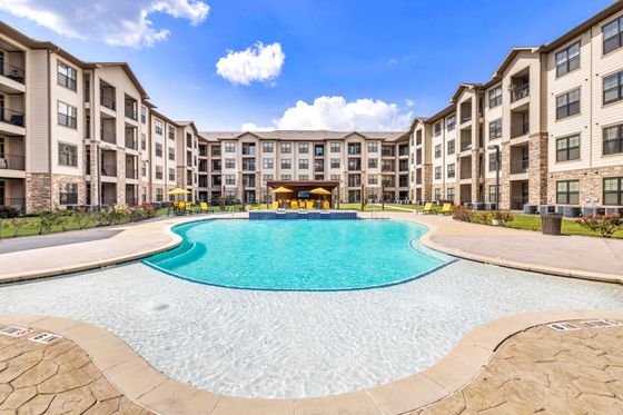 Luxurious apartment complex featuring a stunning resort-style pool with inviting sun loungers and outdoor seating, set against a bright blue sky.