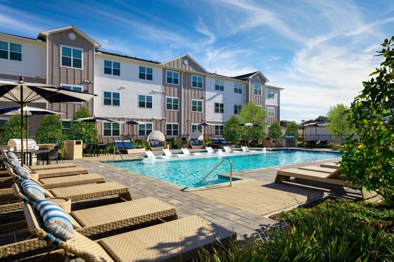 Luxurious apartment complex featuring a sparkling pool with stylish lounge chairs, surrounded by lush greenery and elegant patio seating under vibrant umbrellas.