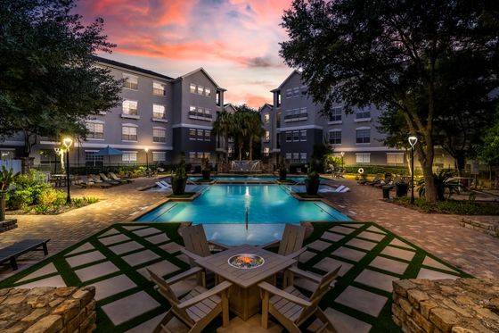 Luxurious apartment exterior featuring a stunning resort-style pool, elegant landscaping, and cozy seating areas under a vibrant sunset sky.