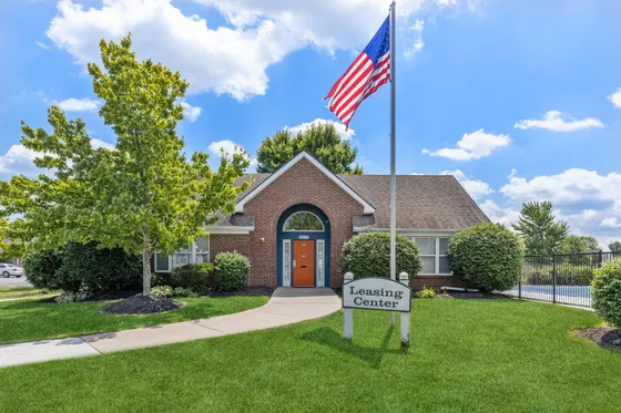 Charming leasing center with a welcoming entrance, lush greenery, and American flag, set under a bright blue sky.