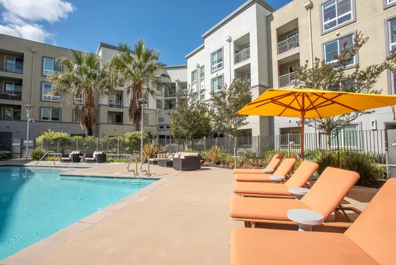 Luxurious apartment complex with a sparkling pool, stylish lounge chairs under vibrant yellow umbrellas, and lush palm trees, perfect for relaxation.