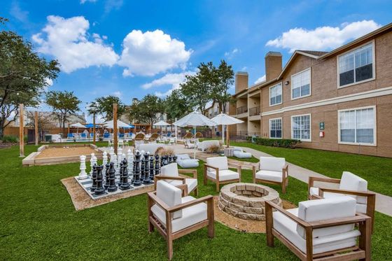 Grassy community area with brick fire pit, cushioned seating, oversized chessboard, umbrellas, and apartment exterior under blue sky.