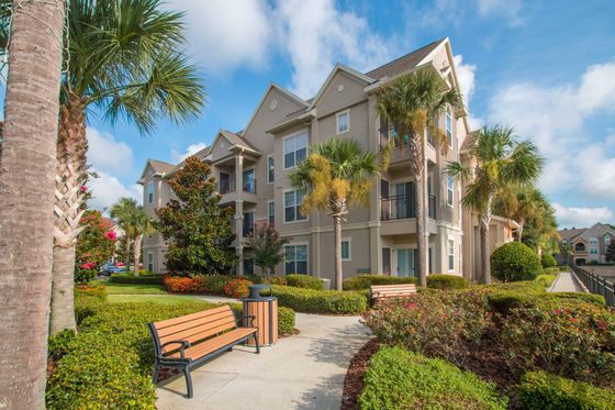 Exterior of a 3-story apartment building with grassy lawn, sidewalk featuring a bench and trash can, palm trees, and native landscaping
