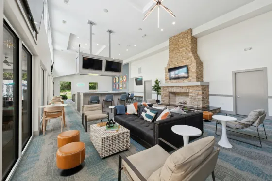 Spacious modern clubhouse lounge with a fireplace, seating area, bar-style counter, wall-mounted TVs, and large windows letting in natural light.