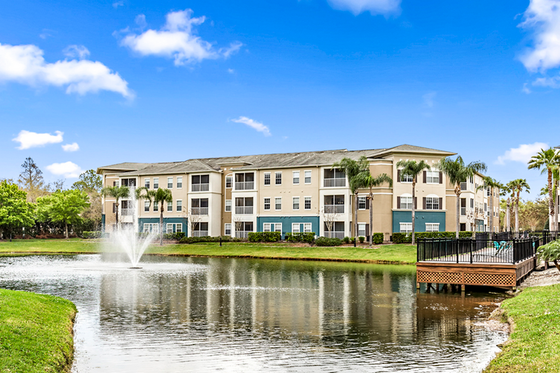 Charming apartment community with palm trees, serene pond, and a beautiful fountain under a clear blue sky.