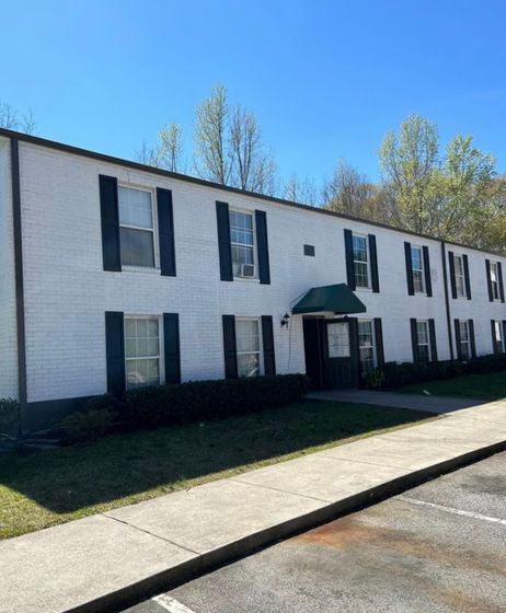 Charming apartment building with white brick exterior and green accents, surrounded by lush greenery and tall trees.