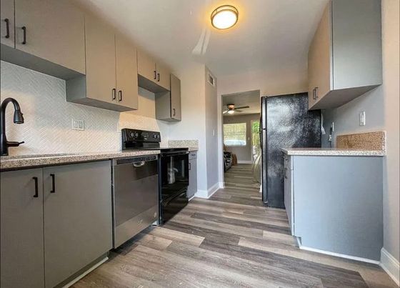 Modern kitchen featuring sleek gray cabinetry, stainless steel appliances, and stylish wood flooring, leading to a bright living area.