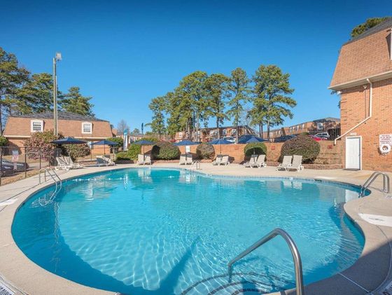 Inviting outdoor swimming pool area with lounge chairs and umbrellas, surrounded by lush greenery and charming brick buildings.
