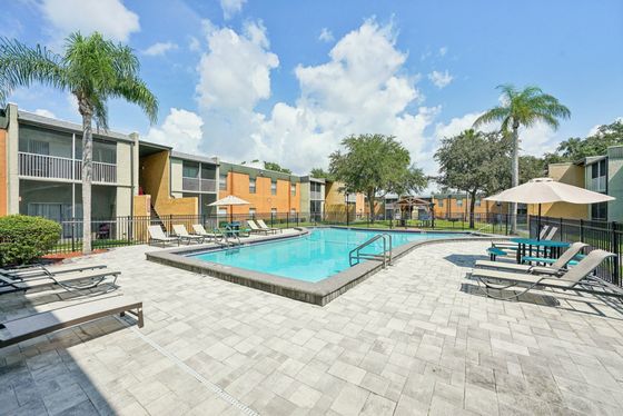 Inviting apartment complex with a pristine pool surrounded by lounge chairs, umbrellas, and lush palm trees under a vibrant blue sky.