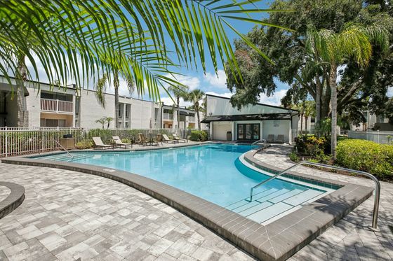 Inviting apartment complex pool with modern design, surrounded by lush greenery and cozy lounge chairs under a bright blue sky.