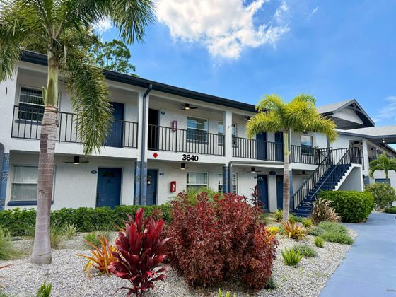 Charming two-story apartment building with tropical landscaping, palm trees, and cozy balconies under a bright blue sky.