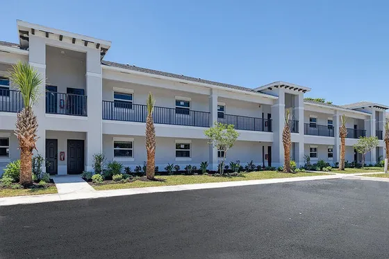 Modern two-story apartment building with balconies, surrounded by lush landscaping and palm trees under a clear blue sky.
