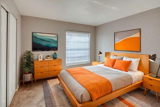 Bright and modern bedroom with stylish furnishings, featuring a comfortable bed with orange accents, natural light from a window, and artistic wall décor.