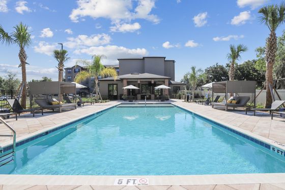 Luxurious apartment complex with a sparkling pool, surrounded by modern lounge chairs, palm trees, and clear blue skies.