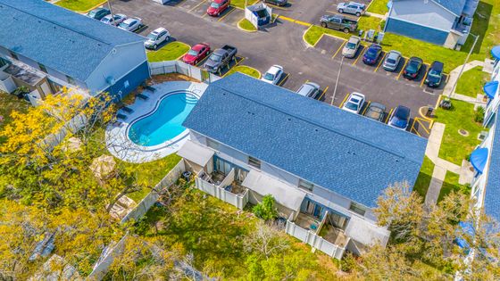 Aerial view of modern apartment complex featuring a sparkling swimming pool and ample parking, surrounded by lush greenery.