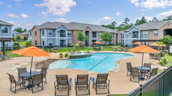 Vibrant apartment complex with a sparkling pool surrounded by lush greenery and modern seating under bright orange umbrellas.