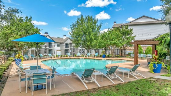 Vibrant apartment complex pool area with comfortable lounge chairs, shaded seating, and lush greenery under a bright blue sky.