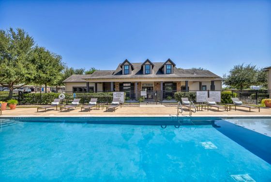 Inviting apartment pool area with sun loungers and a picturesque clubhouse under a clear blue sky.