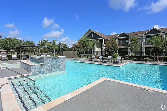 Inviting apartment complex with a sparkling swimming pool, stylish lounge chairs, and lush greenery under a clear blue sky.