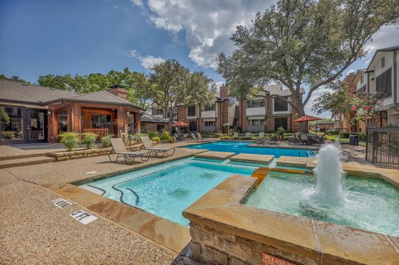 Charming apartment complex featuring a vibrant outdoor pool area with bubbling water features, inviting lounge chairs, and lush landscaping under a bright blue sky.